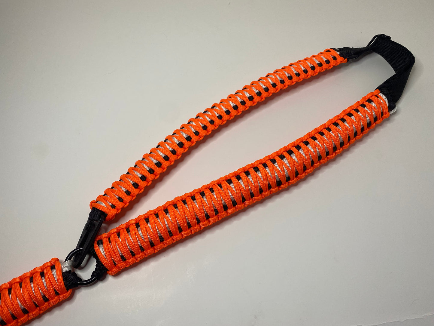a single-point rifle sling made with neon orange, white, and black paracord in the double cobra weave with black hardware.