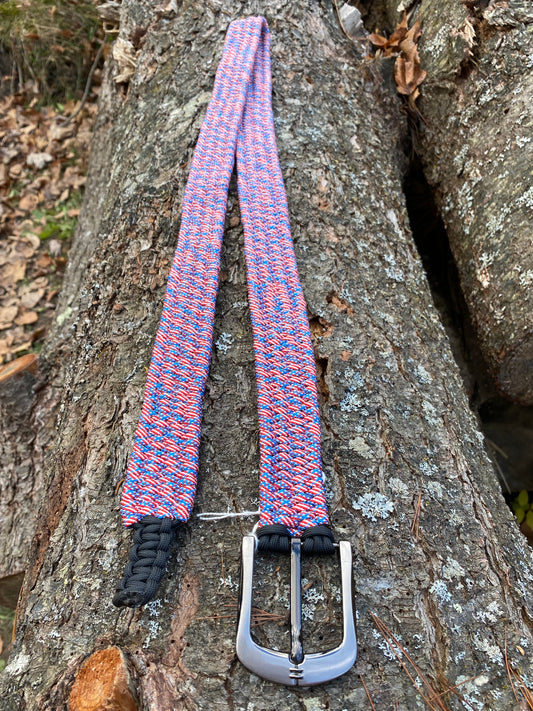 a paracord belt with a large silver buckle made out of USA-flag colored cord sitting on a log.