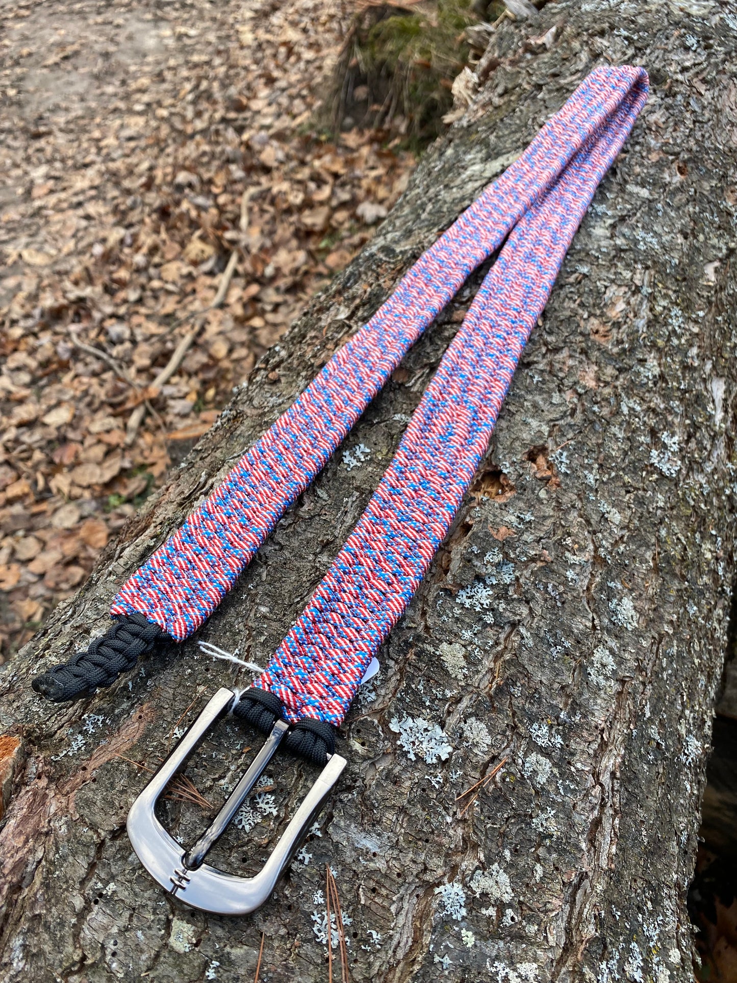 a paracord belt with a large silver buckle made out of USA-flag colored cord sitting on a log.