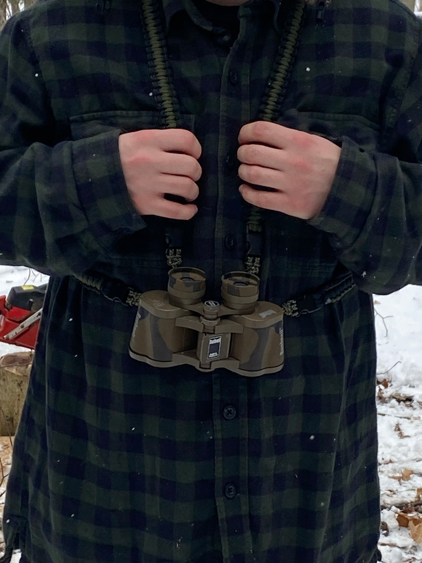 A man wearing a paracord binocular lanyard made with 550 black and olive drab paracord which is carrying a pair of binoculars with a snowy wood in the background