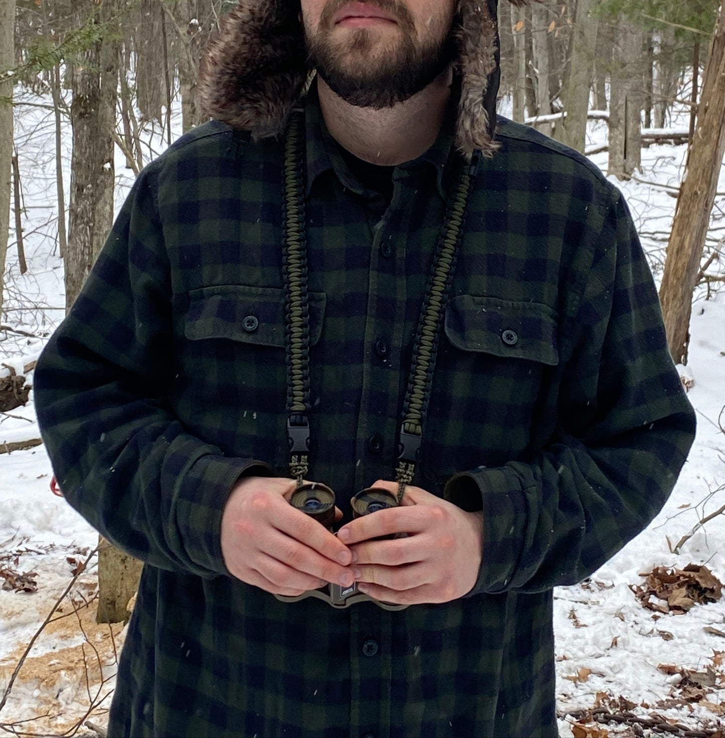 A man wearing a paracord binocular lanyard made with 550 black and olive drab paracord which is carrying a pair of binoculars with a snowy wood in the background